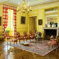 Chambre luxueuse avec des murs jaunes, des meubles ornés, un lustre, une cheminée, des rideaux rouges et blancs et un tapis à fleurs. DR