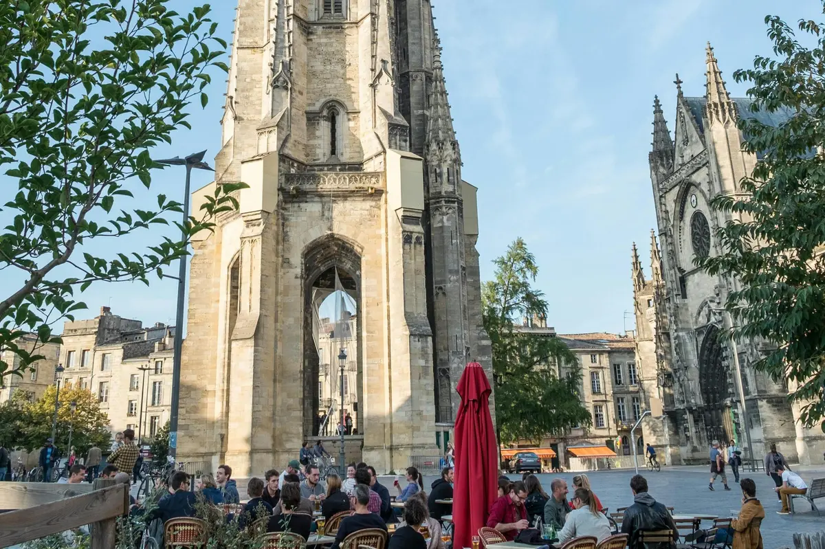 Un café en plein air animé, avec en toile de fond une tour historique en pierre et un ciel bleu clair, où de nombreux convives s'amusent.