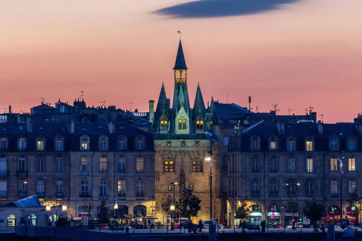 Un bâtiment historique illuminé, doté de tours, à la tombée de la nuit, entouré d'autres bâtiments sous un ciel rose et violet.
