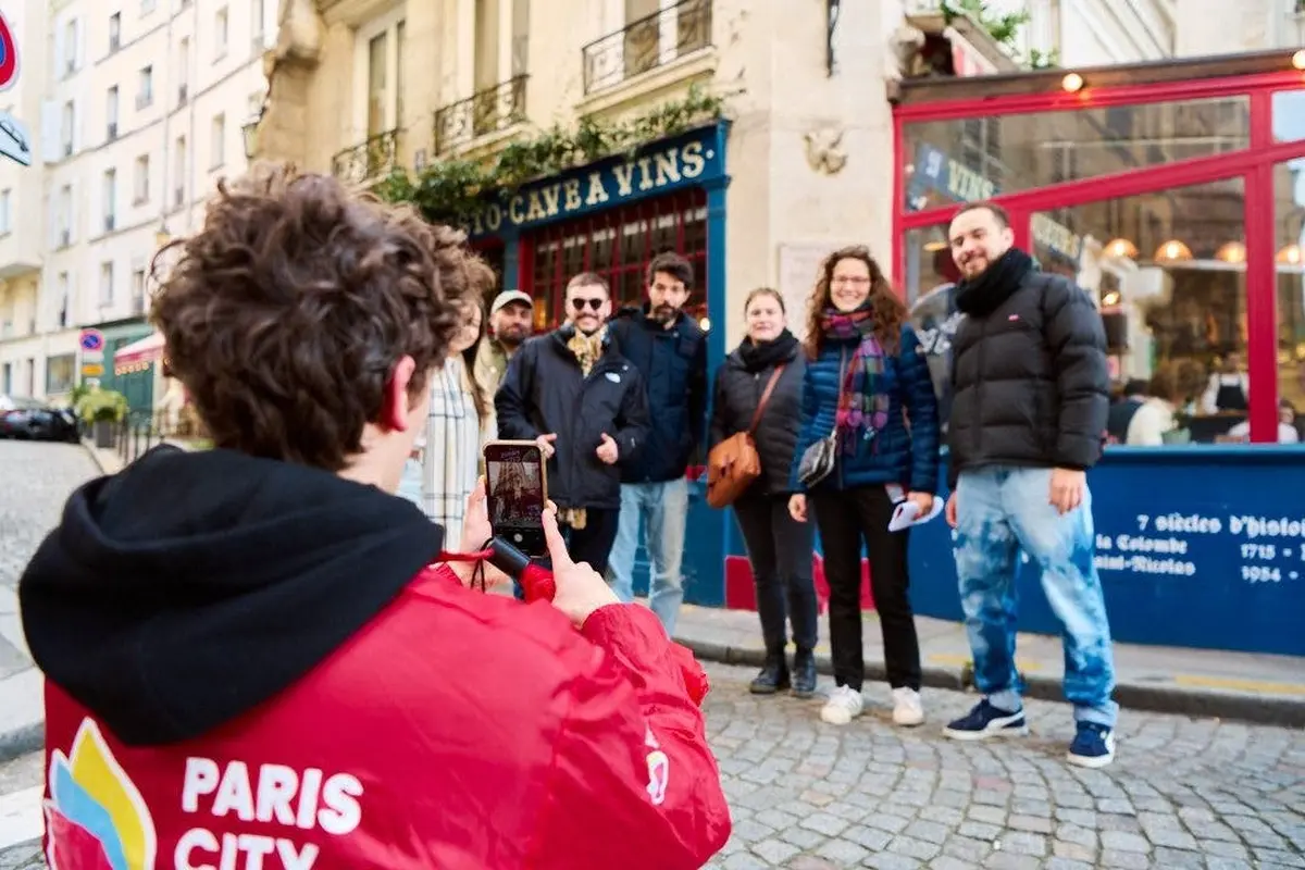 Une personne vêtue d'une veste rouge prend une photo de six personnes posant ensemble devant une devanture bleue dans une rue pavée.