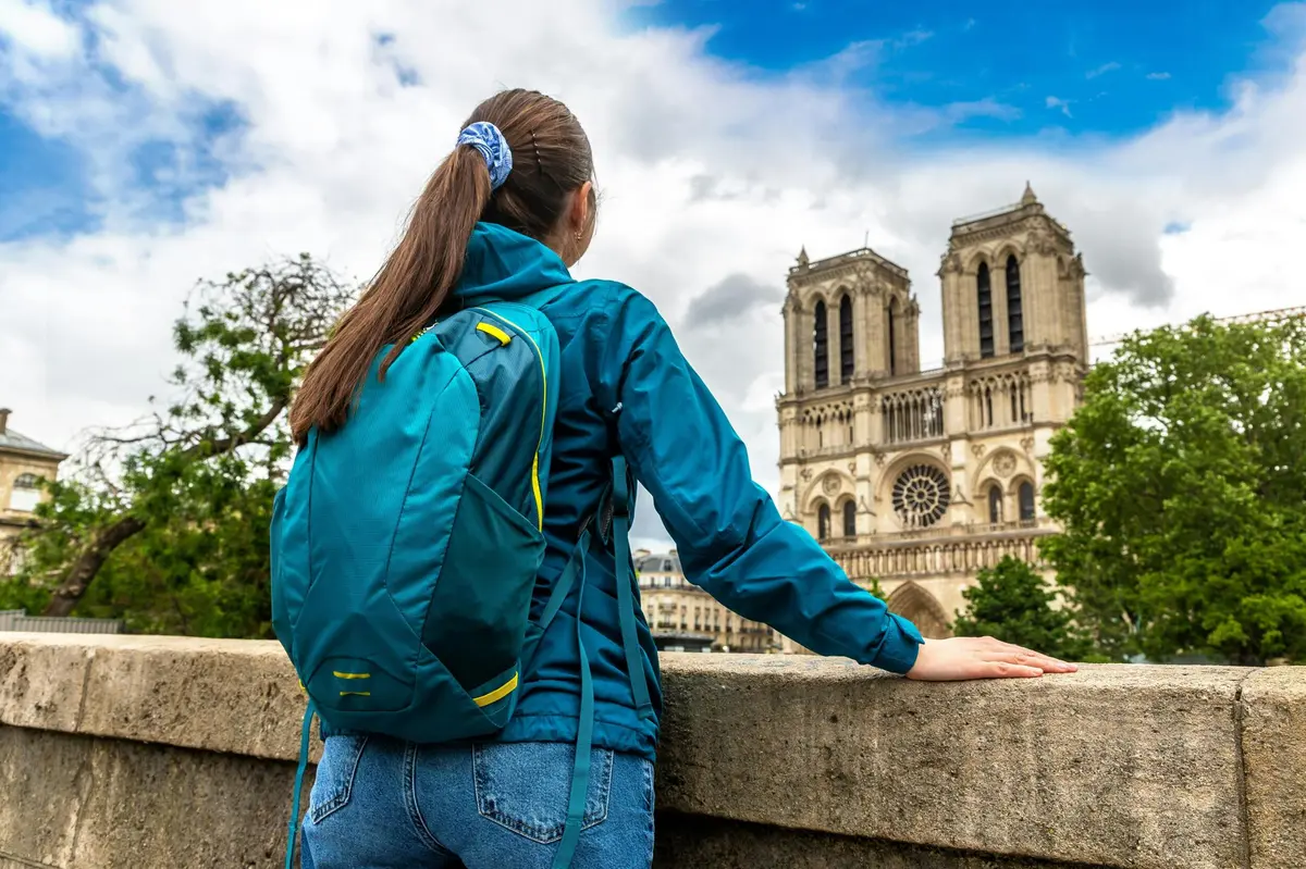 Personne avec un sac à dos et une veste bleus face à la cathédrale Notre-Dame par une journée nuageuse.
