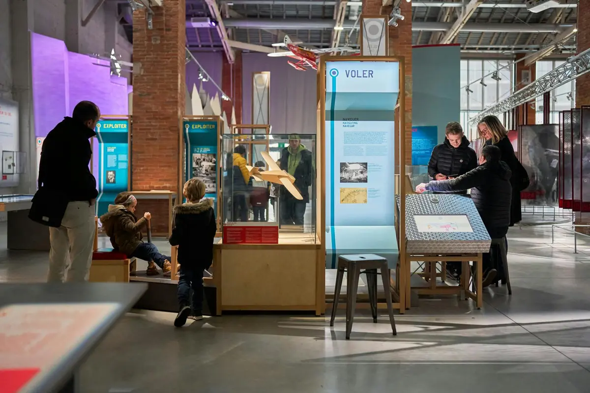 Des visiteurs découvrant les expositions d'un musée, notamment une vitrine intitulée « Voler » et une maquette d'avion exposée dans une vitrine.