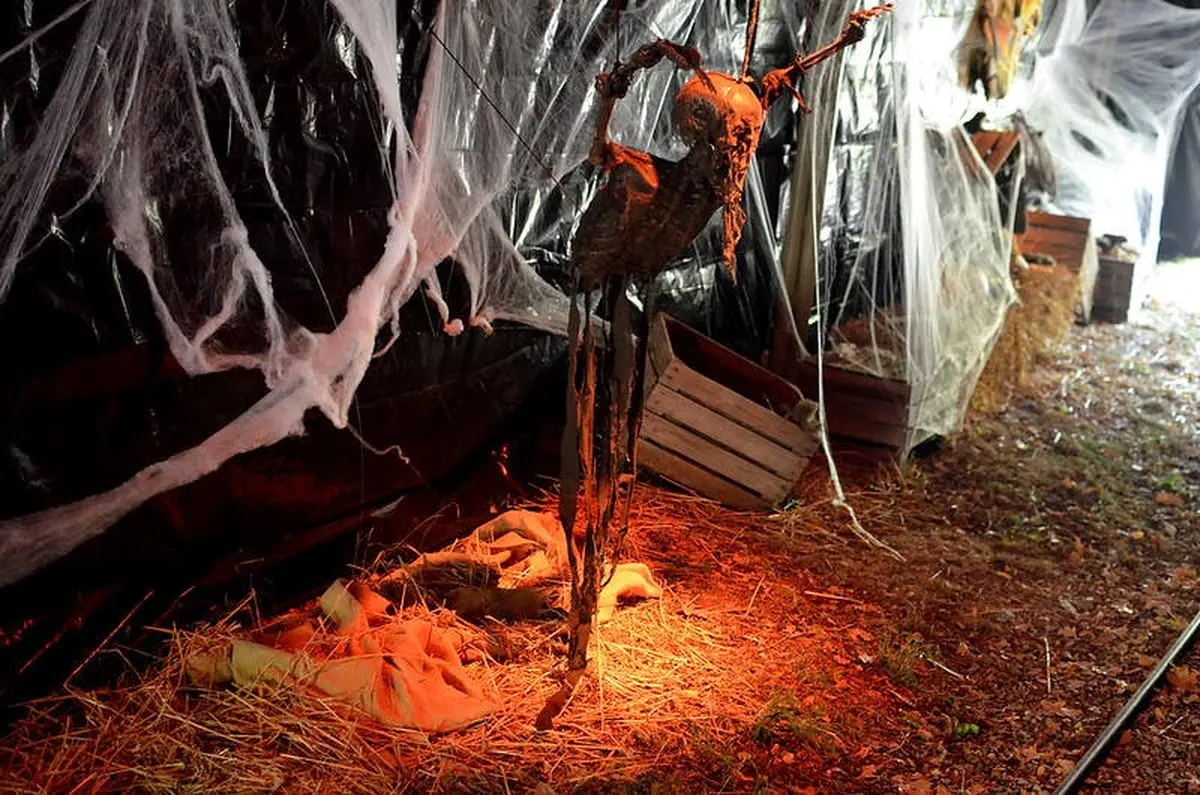 Une figure squelettique effrayante est suspendue à un tissu en lambeaux et à des toiles d'araignée sous un éclairage rouge, entourée de caisses et de toiles de jute sur un sol recouvert de paille.