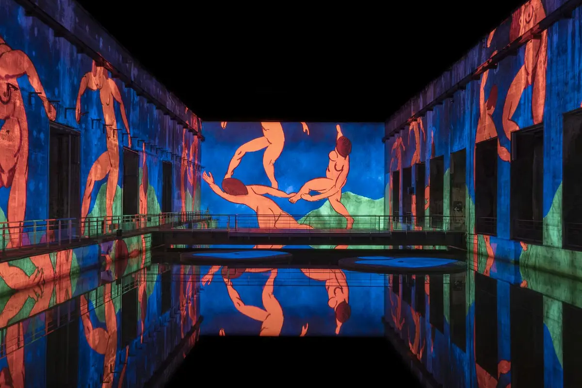 Projection colorée de personnages sur les murs entourant un bassin réfléchissant dans un espace sombre.