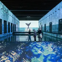 Trois personnes se tiennent dans une grande salle avec des projections numériques d'eau de mer et d'une queue de baleine. &copy; © Cultuirespaces