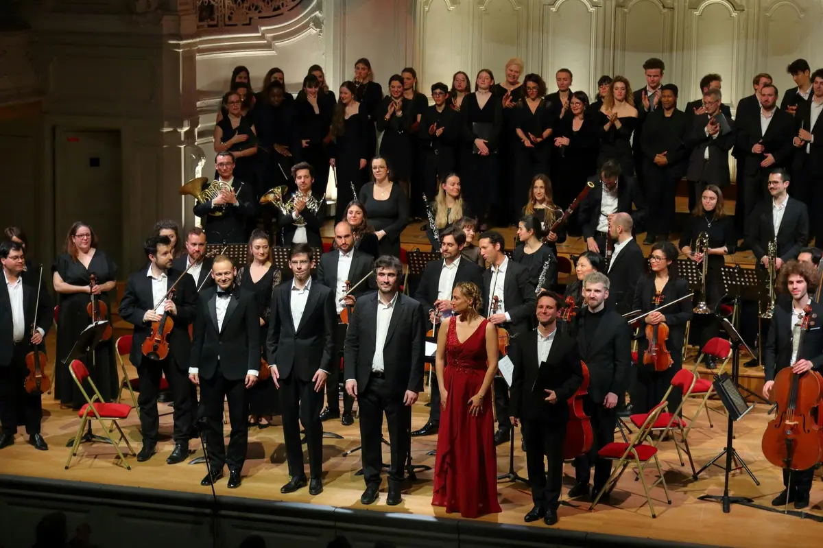 Un orchestre symphonique composé de musiciens et d'une chorale sur scène pendant un spectacle, certains debout et d'autres assis.