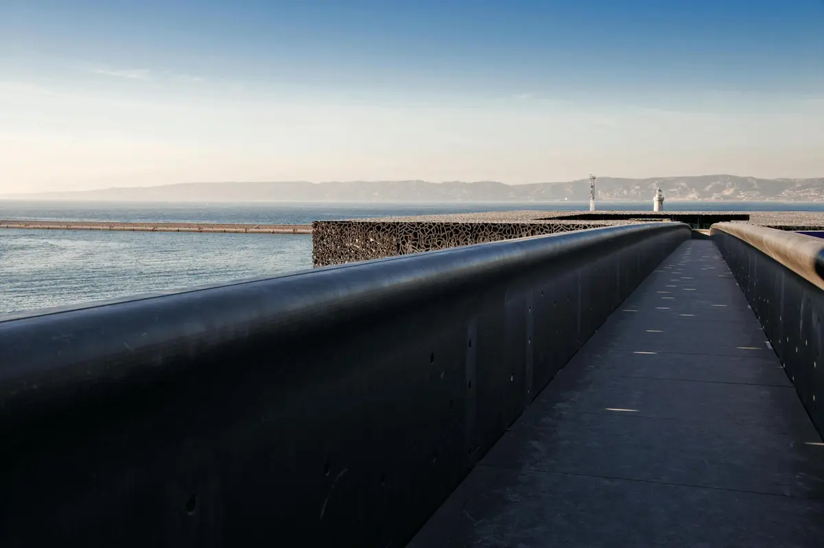 Une promenade en bord de mer avec une balustrade noire, une structure métallique complexe et des montagnes lointaines sous un ciel bleu clair.