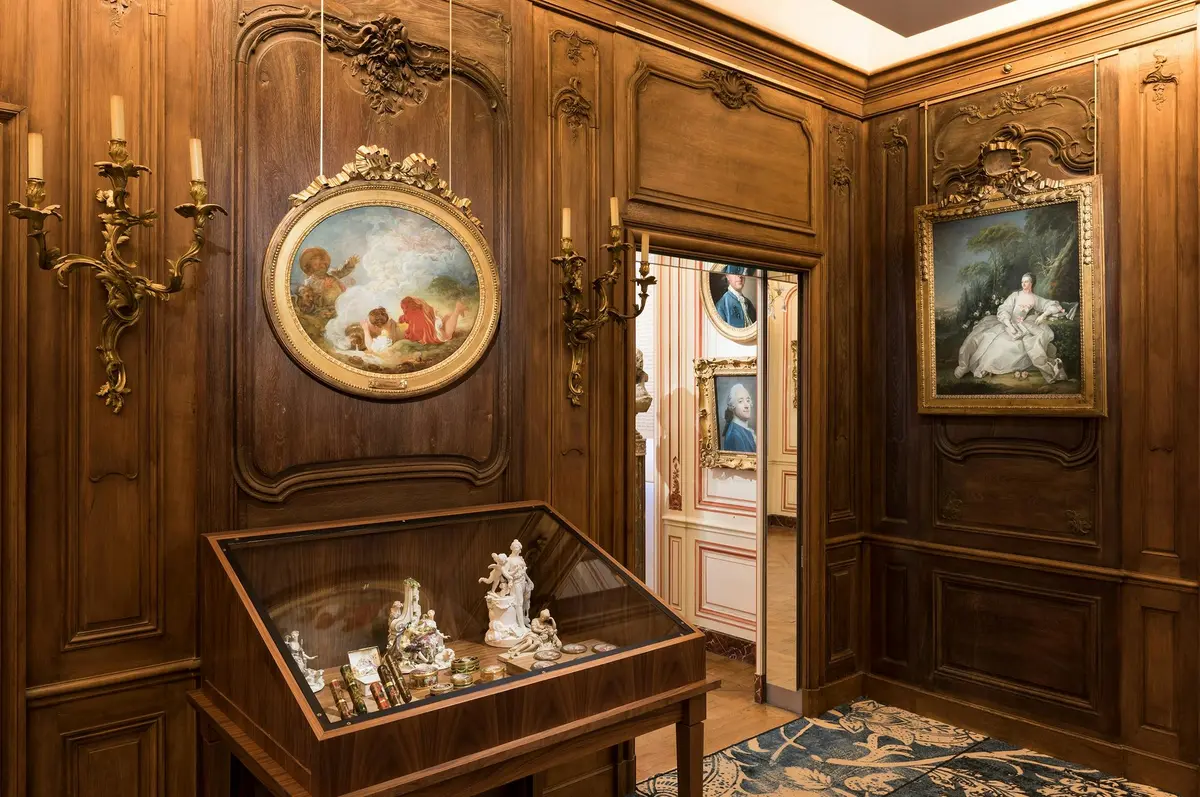 Ornate wooden room displaying figurines in a glass case, with classic paintings and intricate wall moldings. Adjoining room visible.