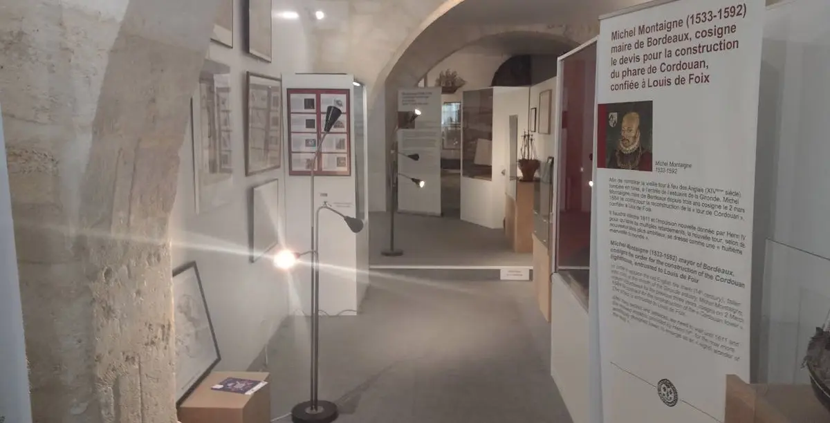 Une exposition muséale comprenant des œuvres d'art encadrées sur les murs, des panneaux d'information, des vitrines et des lampadaires dans un espace lumineux.