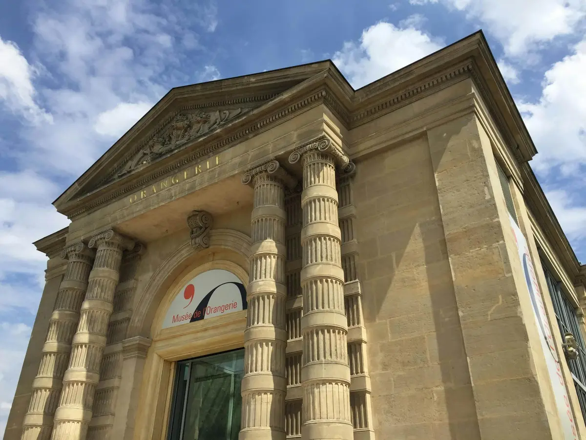 Bâtiment en pierre avec des colonnes ornées et une arche. L'enseigne indique Musée de l'Orangerie. Ciel bleu avec quelques nuages en arrière-plan.