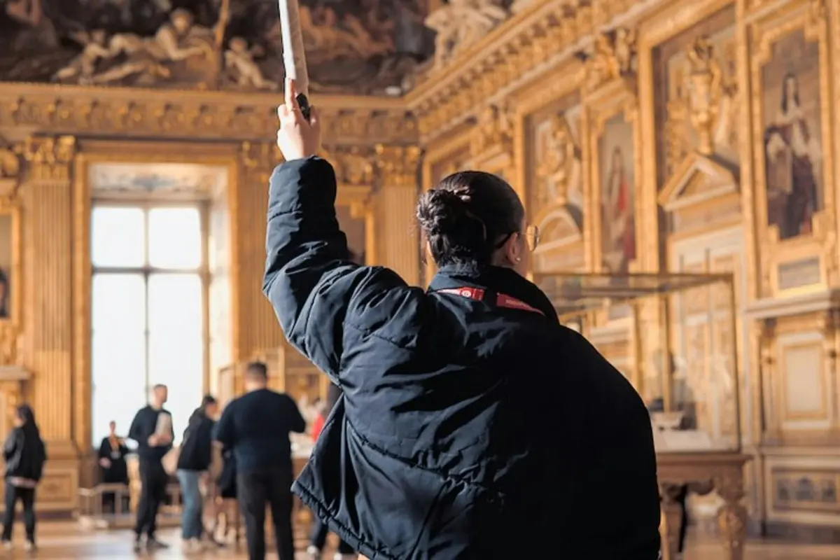 Un guide tenant un bâton dirige un groupe dans une salle ornée et dorée, remplie d'œuvres d'art encadrées et de grandes fenêtres.