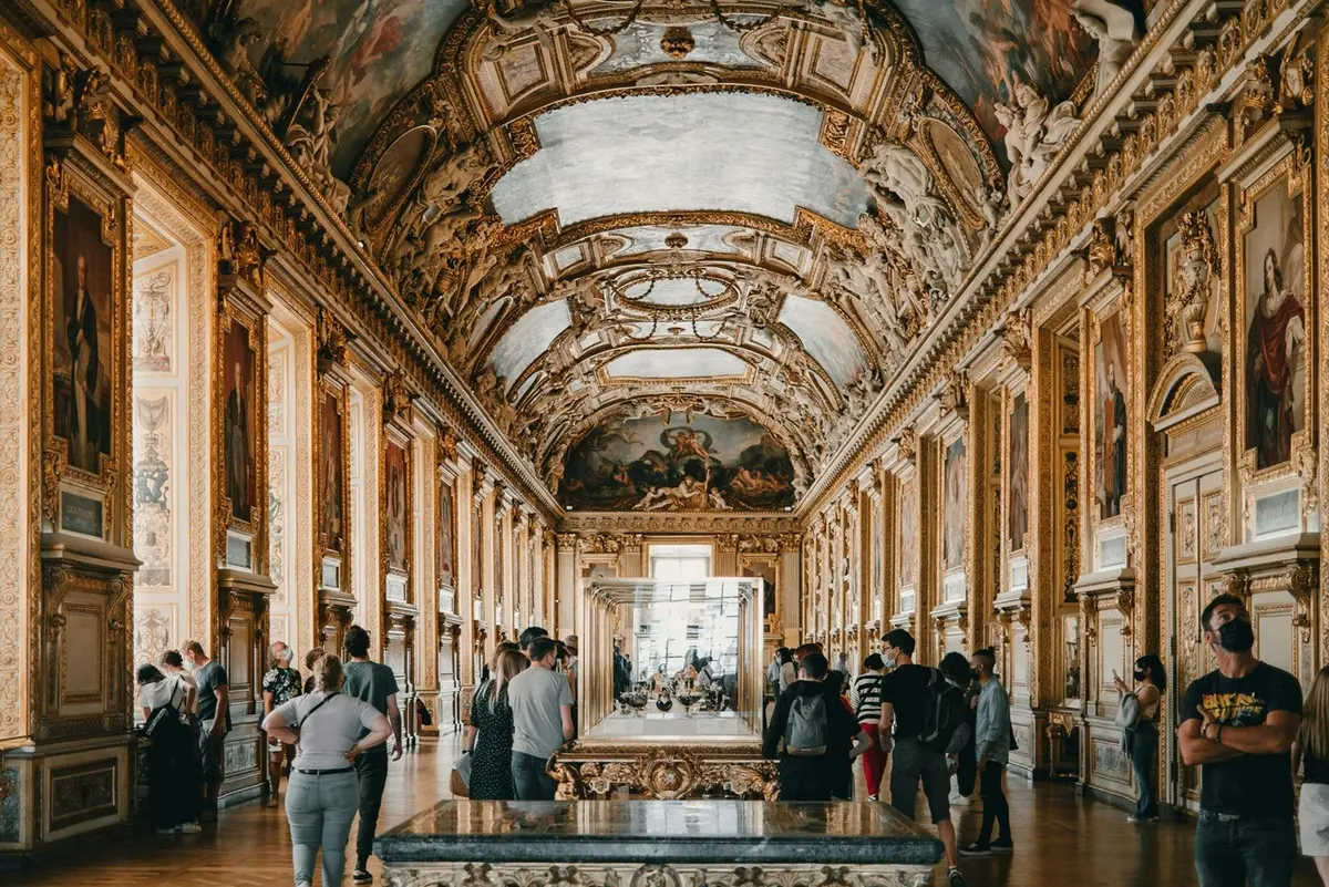 Les gens admirent des œuvres d'art dans une grande galerie aux murs dorés et ornés, au plafond peint et aux vitrines.