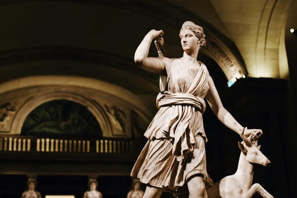 Statue d'une femme drapée tenant un arc, accompagnée d'un cerf, dans un espace intérieur orné.