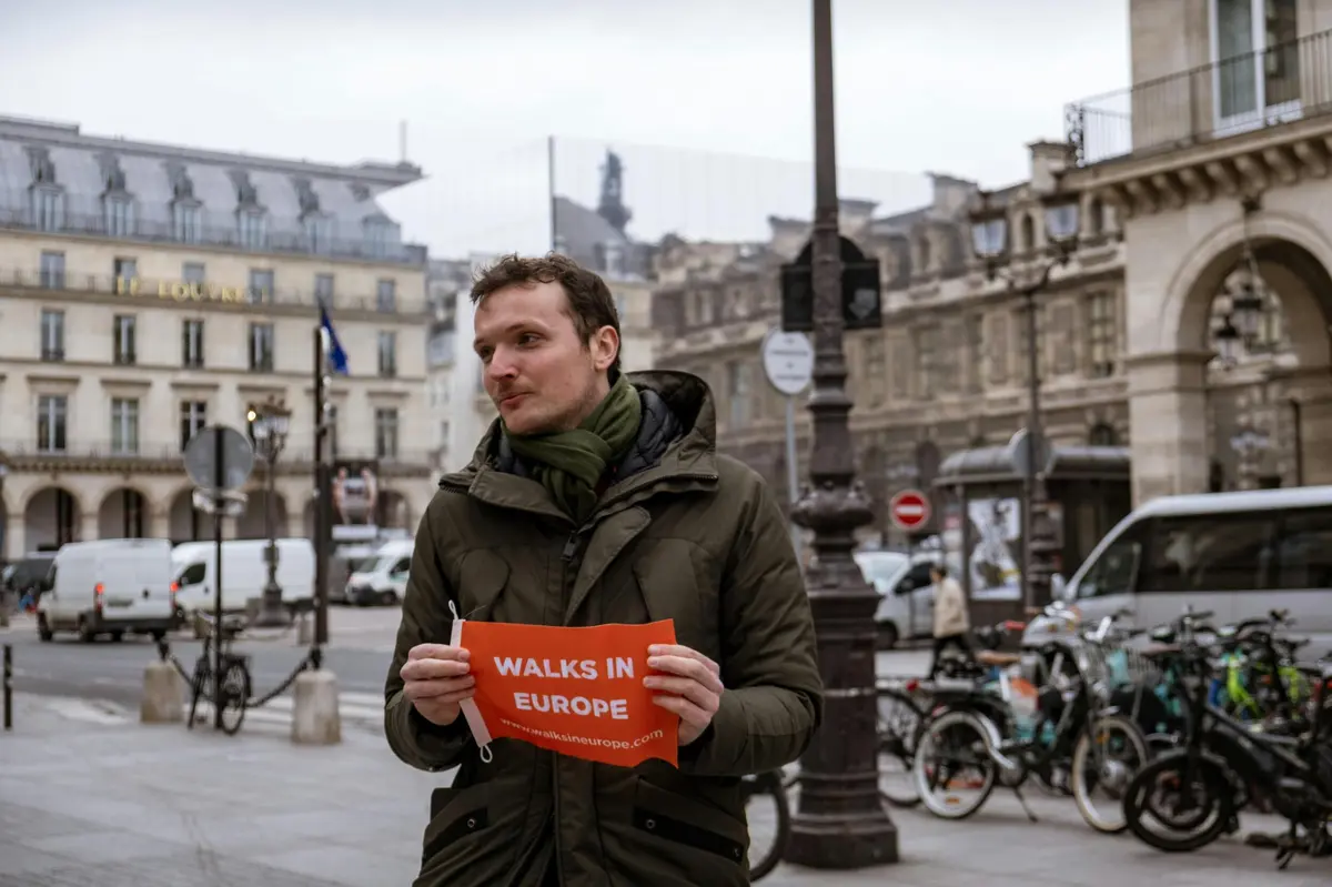 A person in a green coat holding a red sign reading Walks in Europe stands in a European street with buildings and cars.