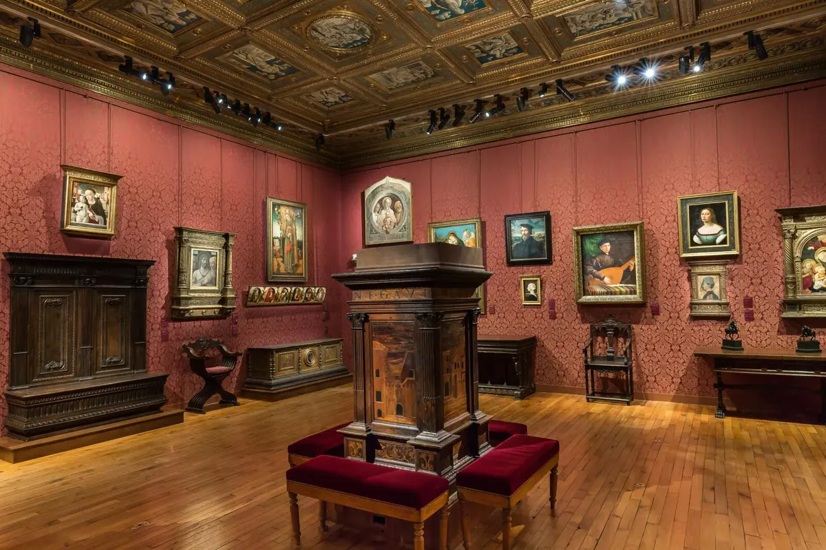 Une galerie d'art aux murs rouges, au sol en bois et au plafond orné. Diverses peintures encadrées et des meubles classiques y sont exposés.