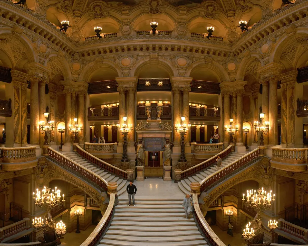 Un grand escalier décoré de lustres et de piliers à l'intérieur d'un bâtiment cossu. Quelques personnes se tiennent sur les marches.