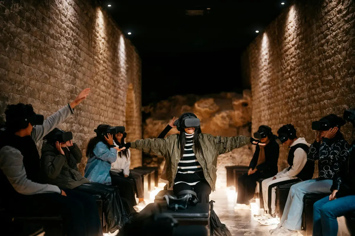 Dans une pièce aux murs de pierre, un groupe de personnes portant des casques de réalité virtuelle est assis sur des bancs, certaines les bras tendus, faisant l'expérience de la réalité virtuelle.