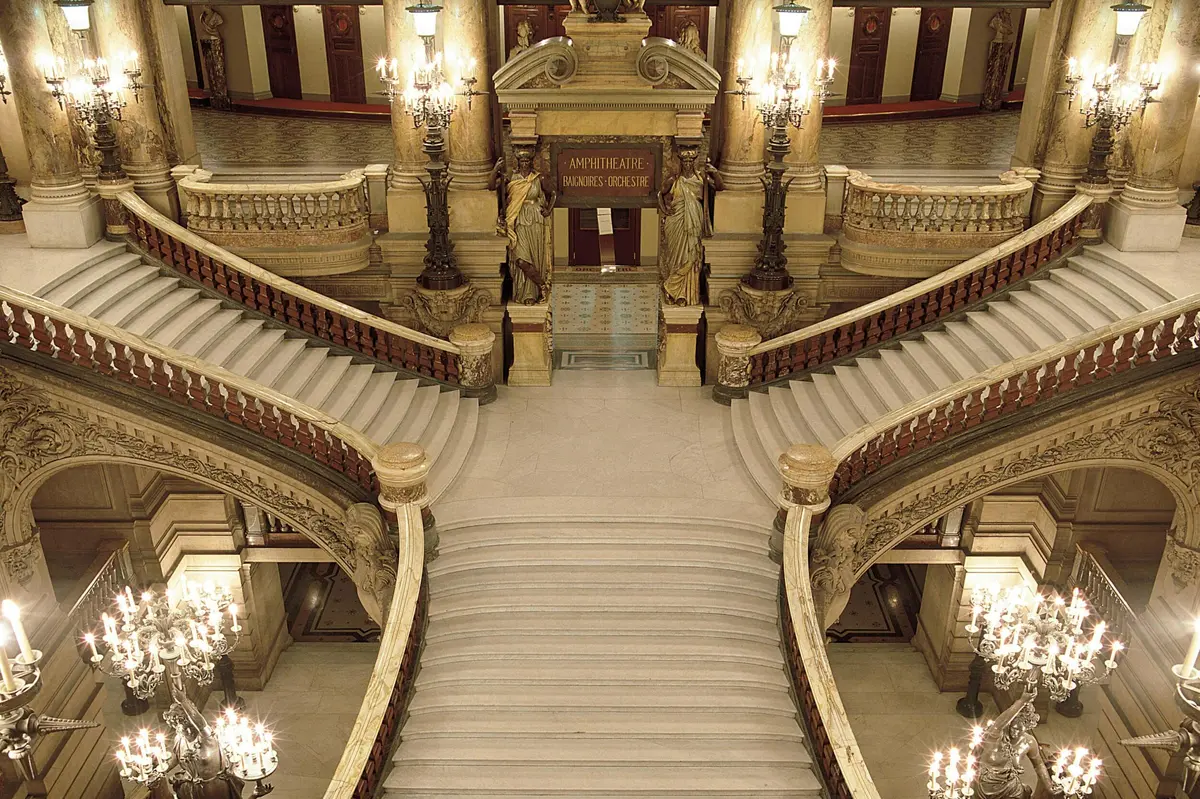 Un grand escalier orné de balustrades sculptées et de statues, menant à une porte portant l'inscription « Amphithéâtre ». Éclairé par des lampes décoratives.