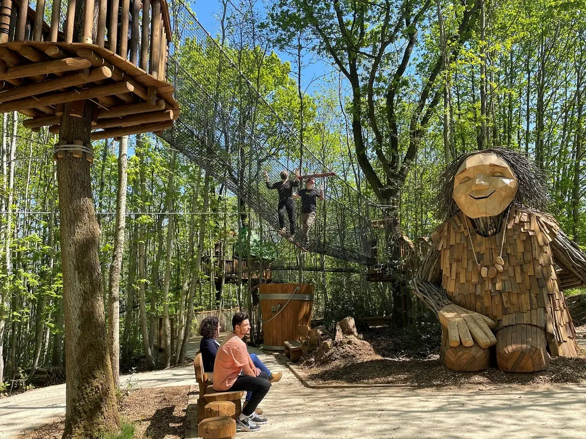 Des personnes sont assises sur des bancs en bois près d'une grande sculpture de troll en bois, tandis que d'autres traversent un pont en filet dans un cadre forestier.