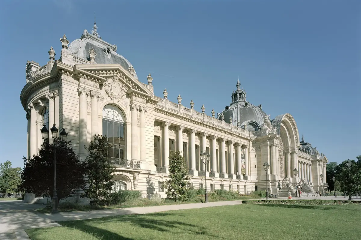 Ornate building with tall windows, columns, and grand domes set in a landscaped garden under a clear blue sky.