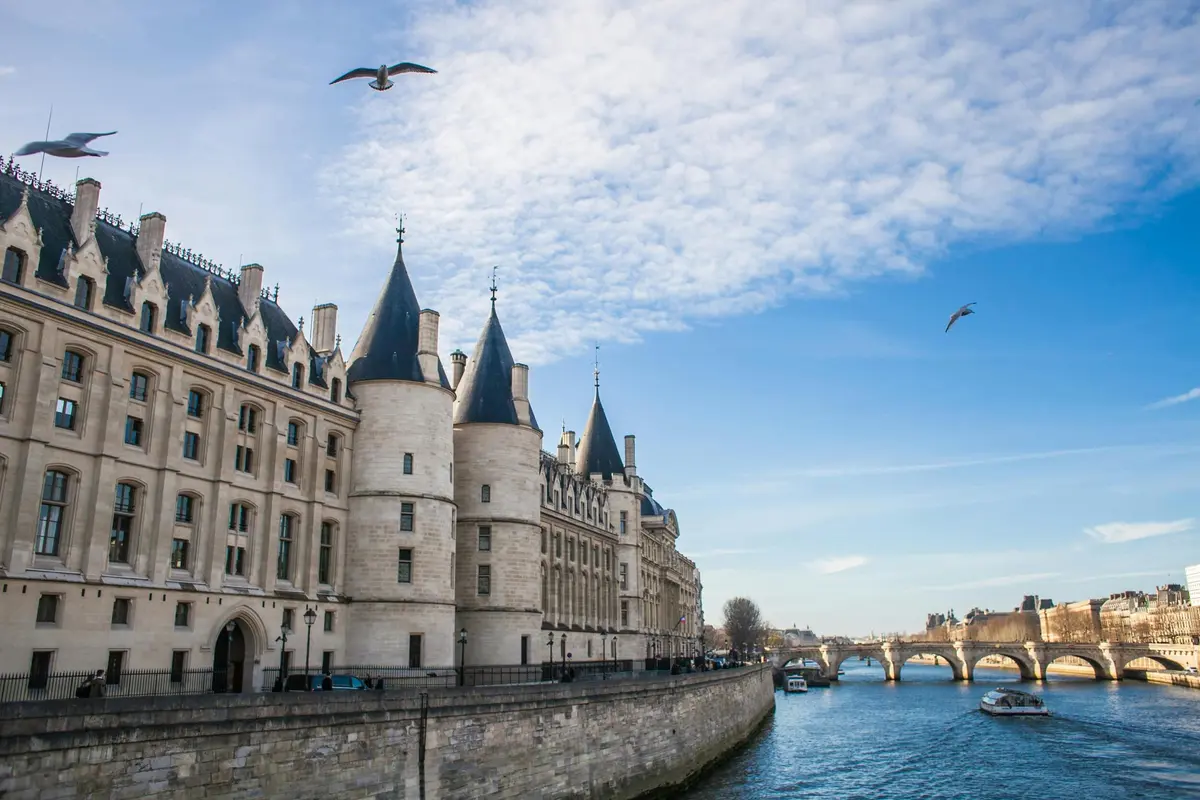 Bâtiment historique situé au bord de la rivière, avec une architecture de style gothique, une promenade au bord de l'eau, des mouettes en vol et un pont au loin.