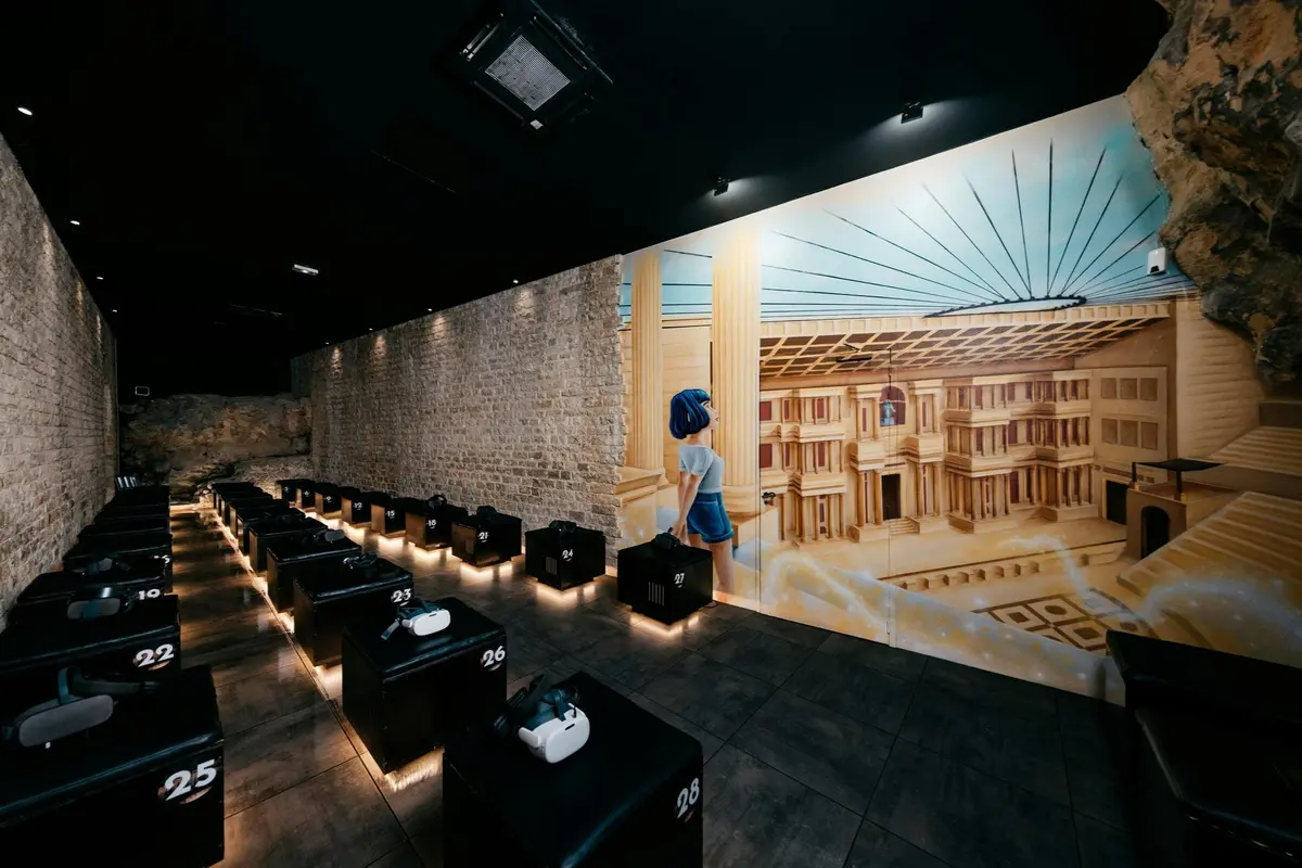 Une pièce ornée d'une fresque représentant un édifice antique, avec des rangées de sièges cubiques noirs équipés de casques de réalité virtuelle, et un mur en briques baigné d'une lumière tamisée.