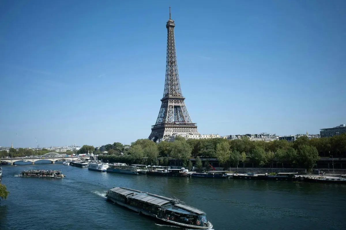 La tour Eiffel vue depuis l'autre rive de la Seine, avec un bateau au premier plan et des arbres bordant la rive.