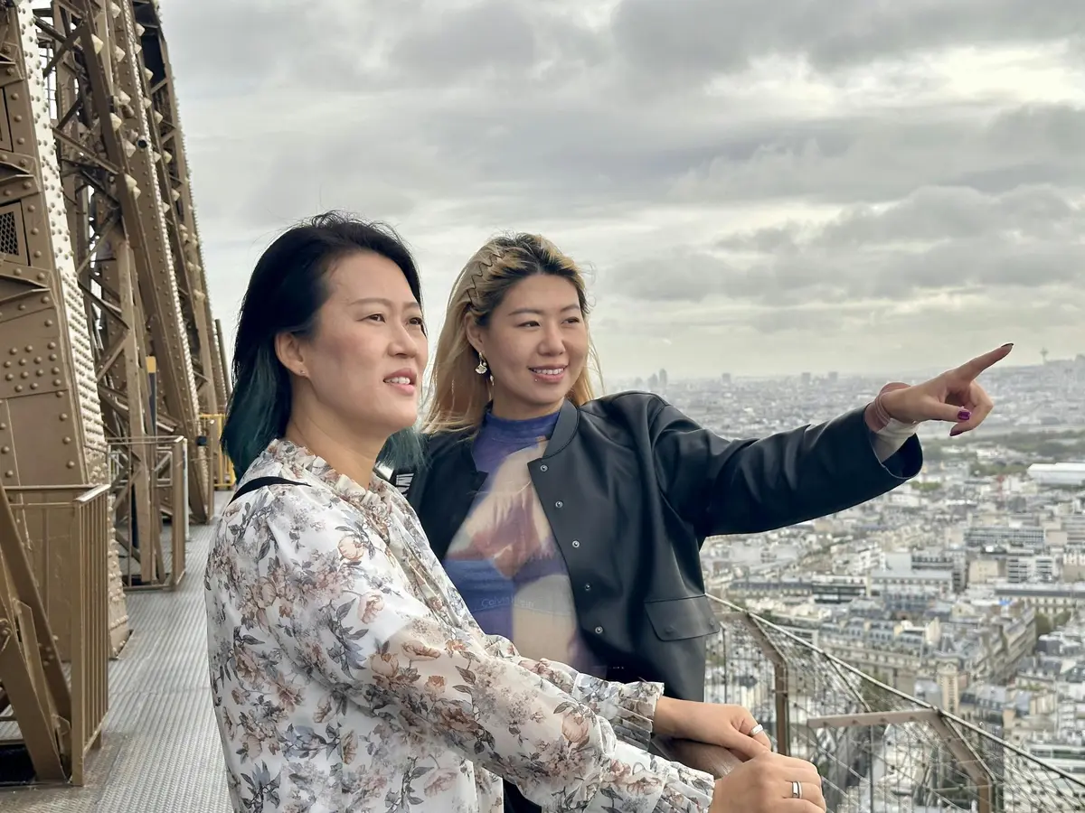 Deux femmes se tiennent sur une plate-forme métallique, près d'une balustrade sur un pont d'observation, avec un paysage urbain visible à l'arrière-plan.