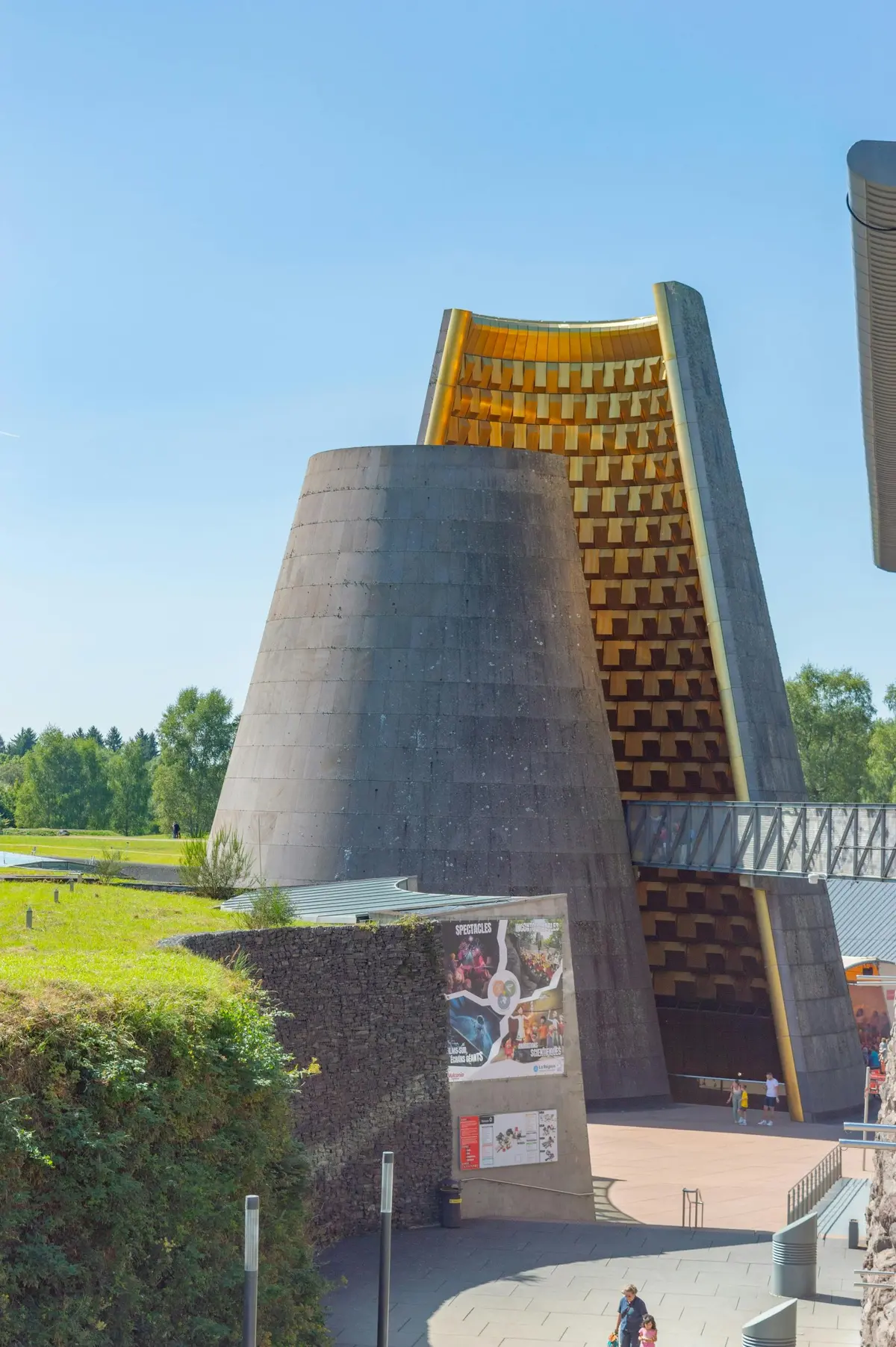 Un grand bâtiment moderne en forme de cône avec une allée adjacente, un environnement herbeux et une affiche près de l'entrée.