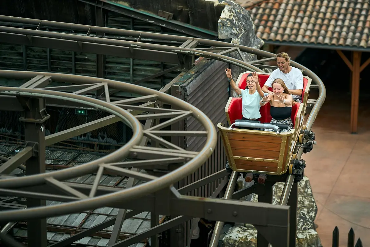 Trois personnes montent sur des montagnes russes avec des expressions d'excitation, en passant devant des rochers et des bâtiments.