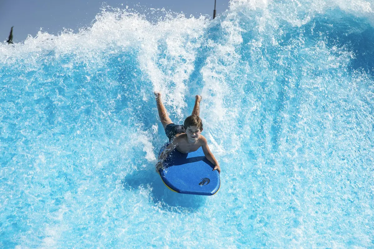 Dans un parc aquatique, une personne descend une vague sur un bodyboard, l'eau bleue éclaboussant autour d'elle.