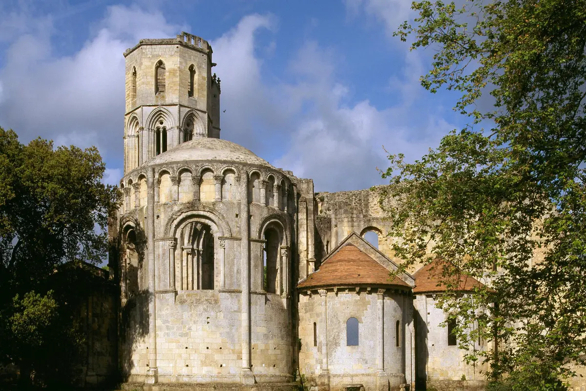 Ancien bâtiment en pierre avec des fenêtres cintrées et une tour cylindrique, entouré d'arbres sous un ciel partiellement nuageux.