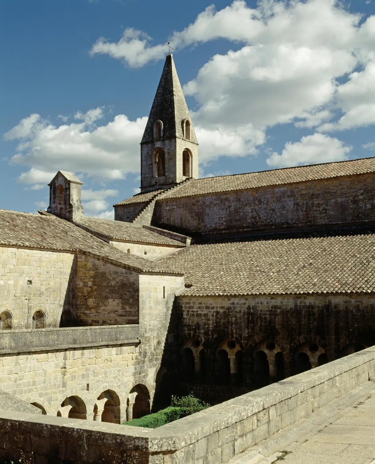 Des bâtiments en pierre avec des cloîtres voûtés et un haut clocher sous un ciel partiellement nuageux.