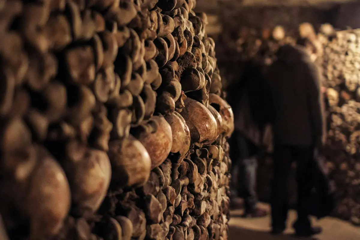 Un mur constitué de crânes et d'os humains empilés, dans un espace souterrain faiblement éclairé, avec des silhouettes floues en arrière-plan.
