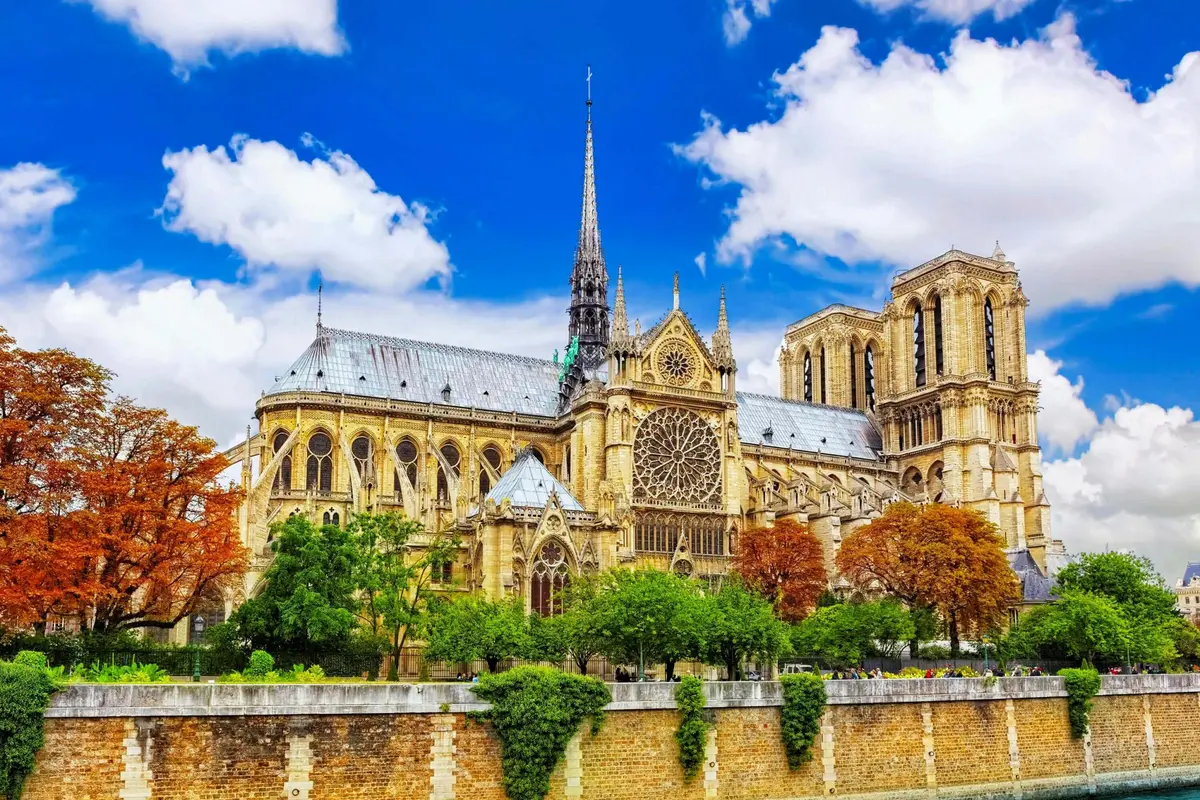 La cathédrale Notre-Dame de Paris, avec son architecture gothique et ses sculptures en pierre détaillées, entourée d'arbres sous un ciel bleu.