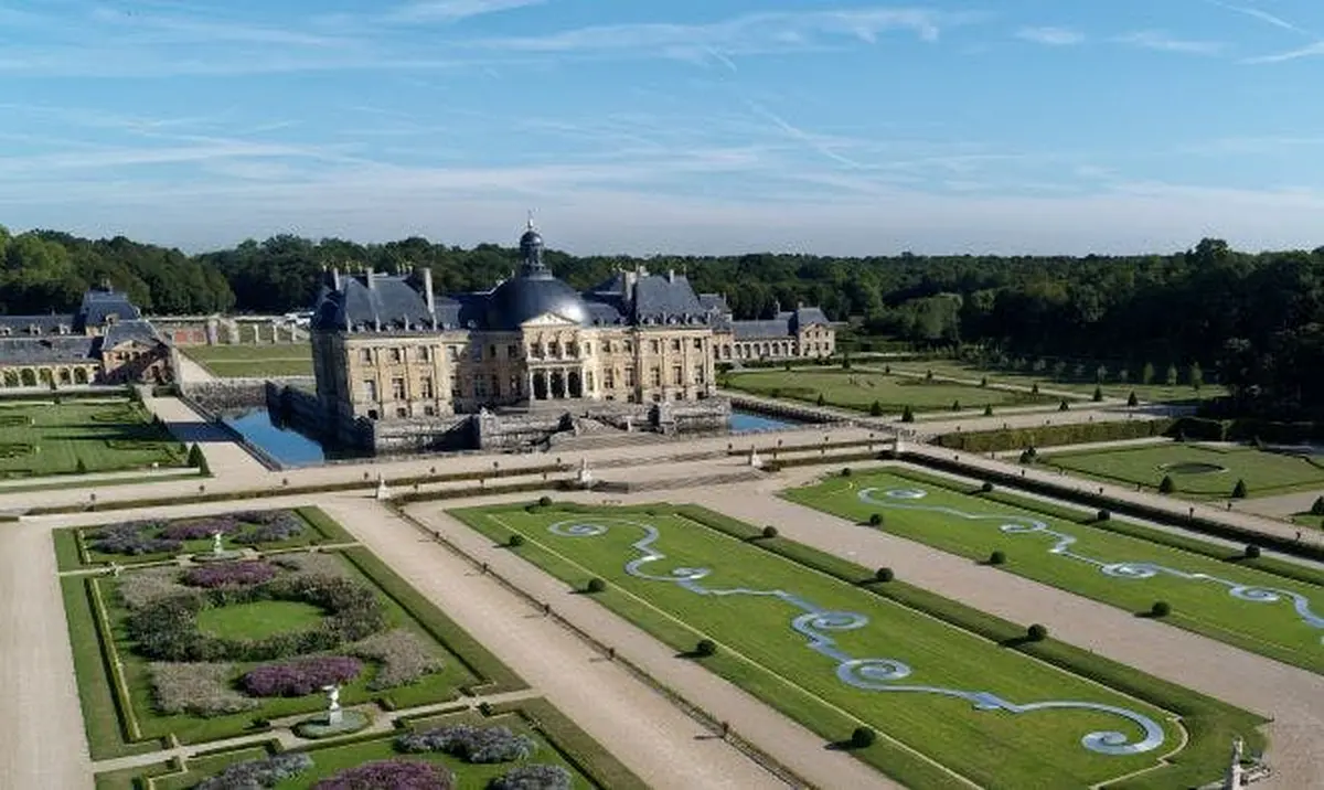 A large historic château surrounded by formal gardens with symmetrical paths, lawns, and water features under a blue sky.