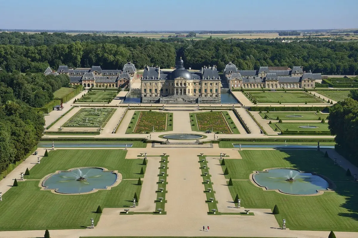 Vue aérienne d'un grand domaine avec un palais central, des jardins symétriques, des fontaines et des arbres environnants.