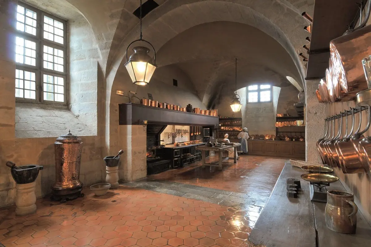 Une cuisine historique avec des plafonds voûtés en pierre, des ustensiles de cuisine en cuivre et une personne travaillant au comptoir à l'arrière-plan.