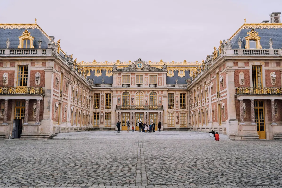 Visitors stand and walk in the expansive courtyard of an ornate palace with symmetrical architecture and gilded details.