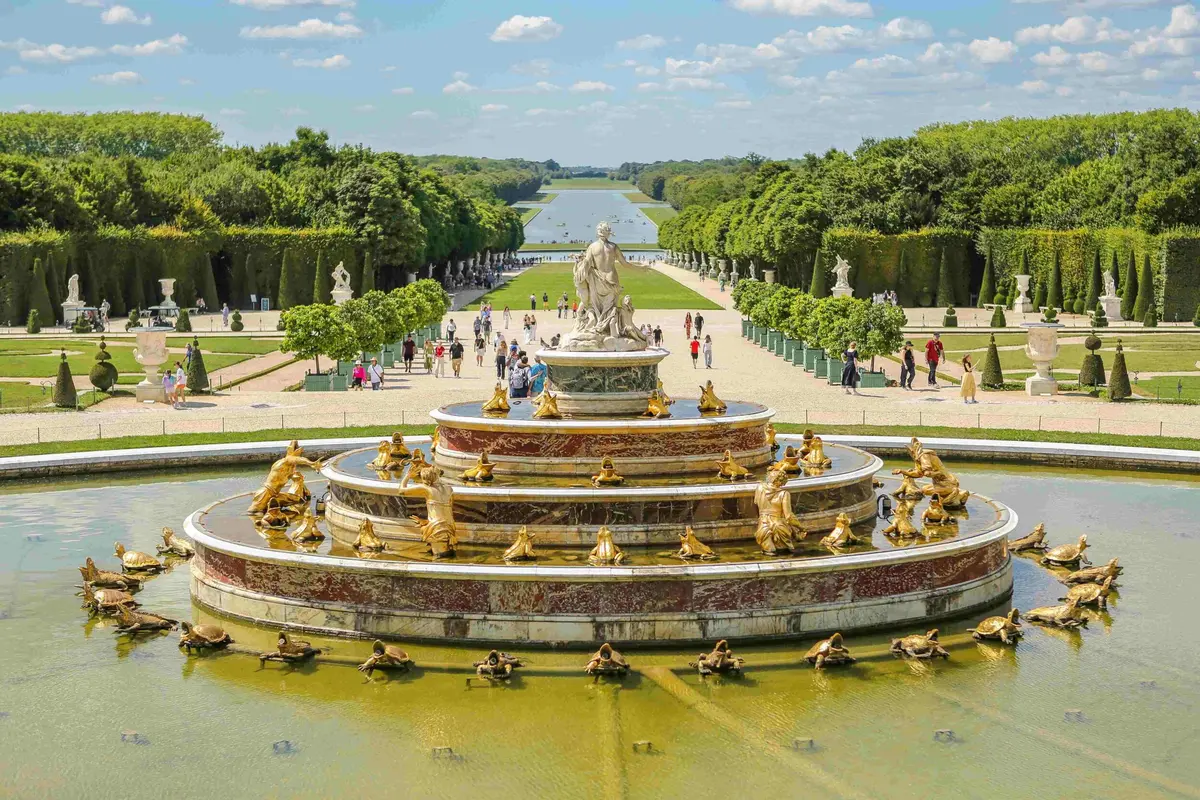 Grande fontaine ornée de statues dorées, entourée de jardins soignés et d'allées où se promènent des gens, sous un ciel bleu.