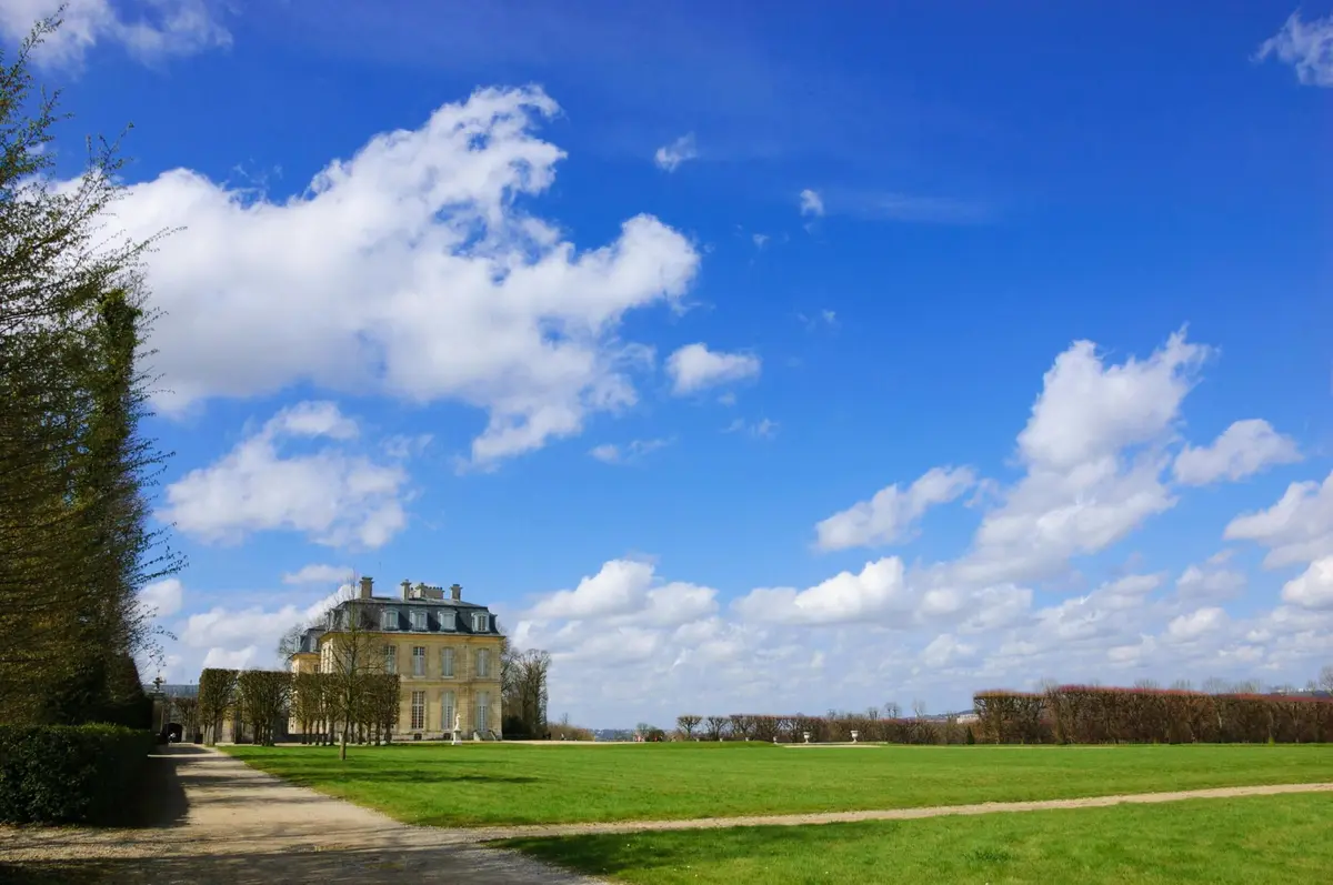Un grand bâtiment de deux étages avec un toit mansardé se trouve à côté d'une vaste pelouse verte sous un ciel bleu vif avec des nuages épars.