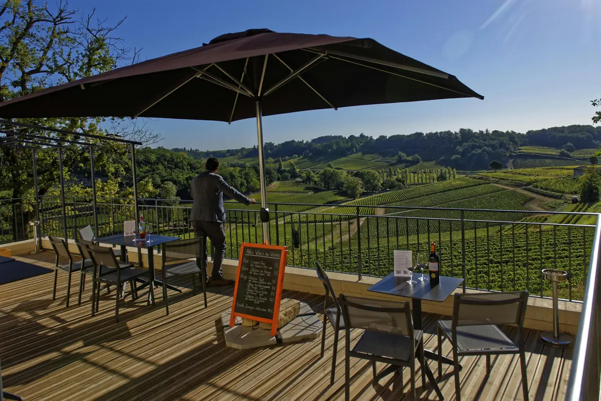 Terrasse d'un restaurant en plein air avec des tables et des chaises sous de grands parasols, avec vue sur un vignoble luxuriant. Une personne se tient près de la balustrade.
