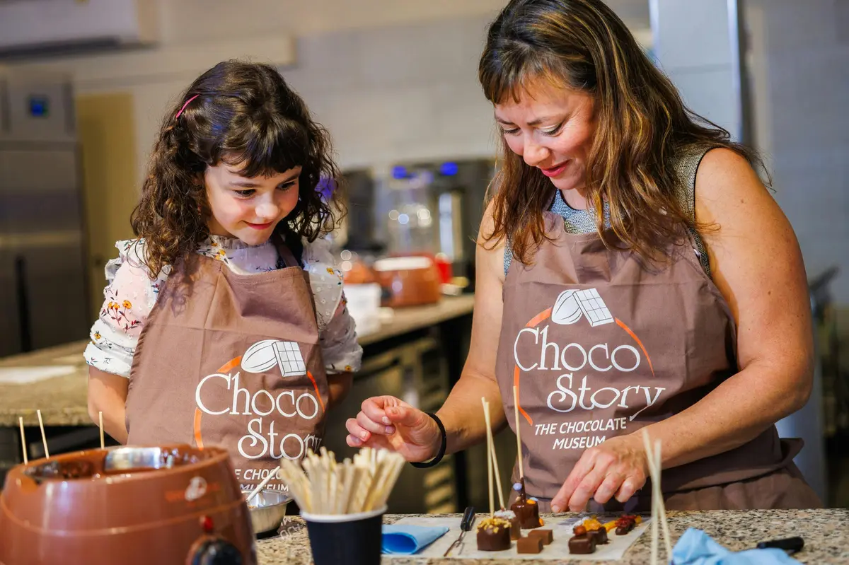 Deux personnes portant des tabliers étiquetés Choco Story décorent des friandises au chocolat sur des bâtonnets dans une cuisine.