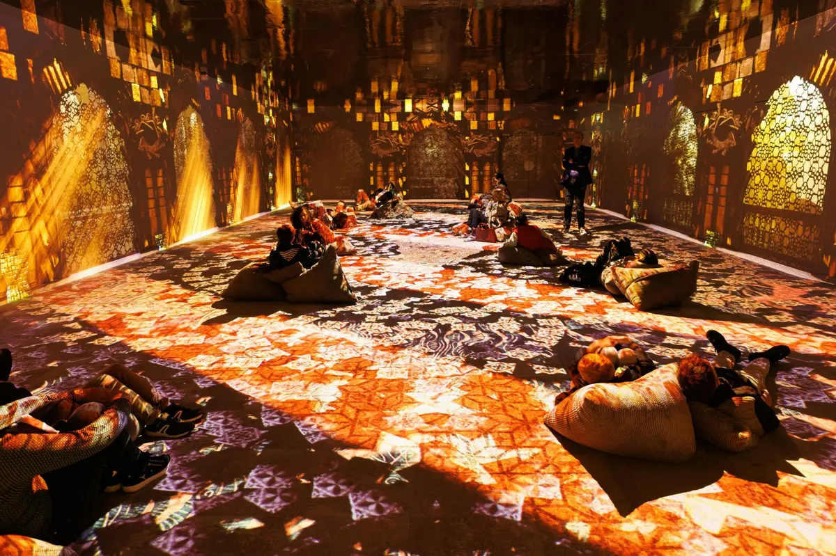 Une salle avec des projections numériques immersives sur les murs et le sol, représentant des motifs vibrants, et des personnes assises sur des coussins.