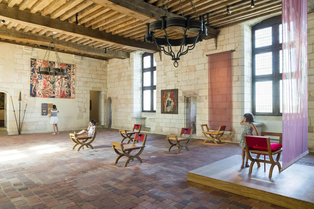 Une salle au décor médiéval avec des peintures, des chaises en bois avec des coussins rouges, un sol en briques et trois personnes qui observent les œuvres d'art.