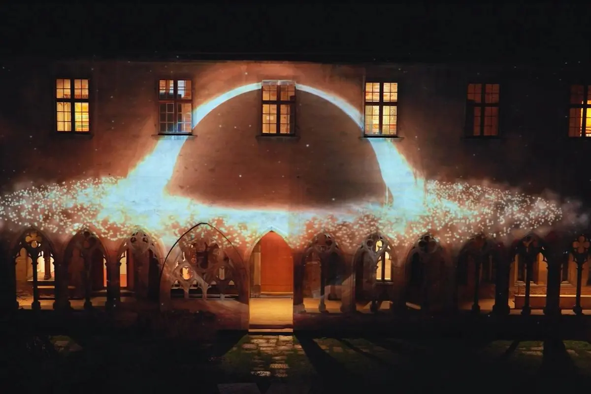 Vue nocturne d'un bâtiment aux fenêtres cintrées, éclairé par une projection lumineuse formant un arc lumineux et des particules.