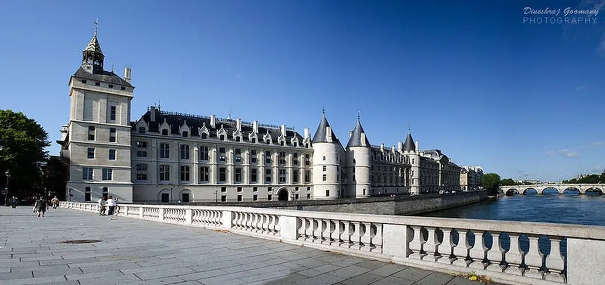 Un palais historique avec des tours pointues et des rangées de fenêtres, vu depuis une balustrade en pierre sous un ciel bleu clair.