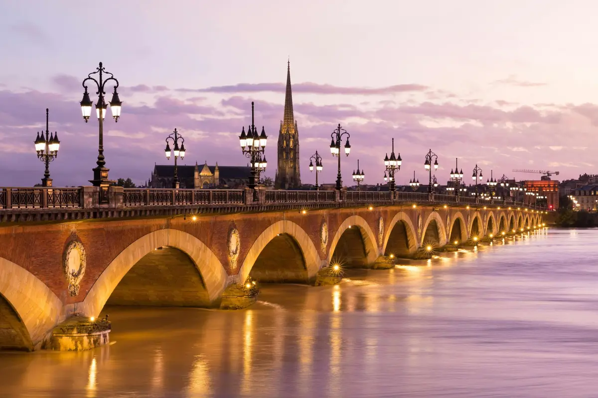 Un pont en arc illuminé enjambe une rivière calme au crépuscule, avec des lampadaires et une haute flèche d'église en arrière-plan.