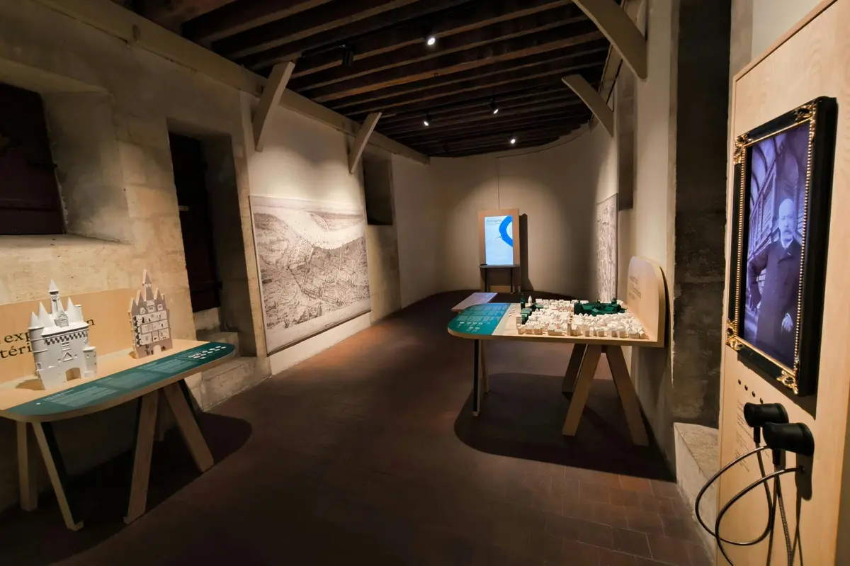 Une salle d'exposition faiblement éclairée, avec des tables en bois sur lesquelles sont exposées des maquettes architecturales blanches et vertes, ainsi que des croquis accrochés aux murs.