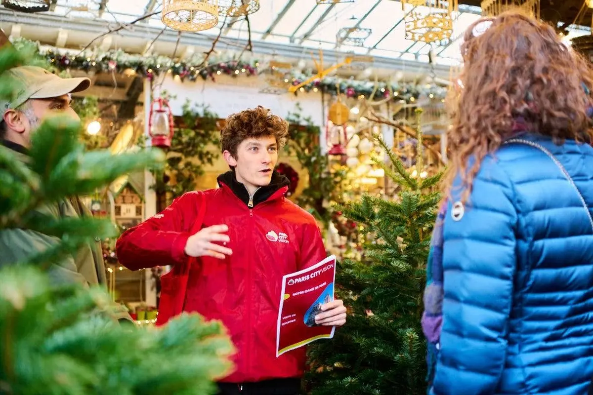 Un homme en veste rouge tient une brochure et parle à un autre homme au milieu de décorations et d'arbres de Noël dans un décor intérieur festif.